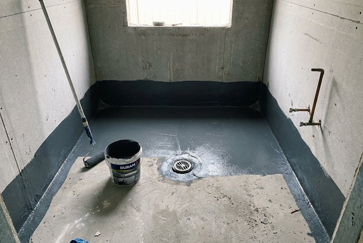 Charcoal waterproofing membrane rolled onto the floor and up the walls of a bathroom mid-renovation, with the floor waste detailed, before tiling