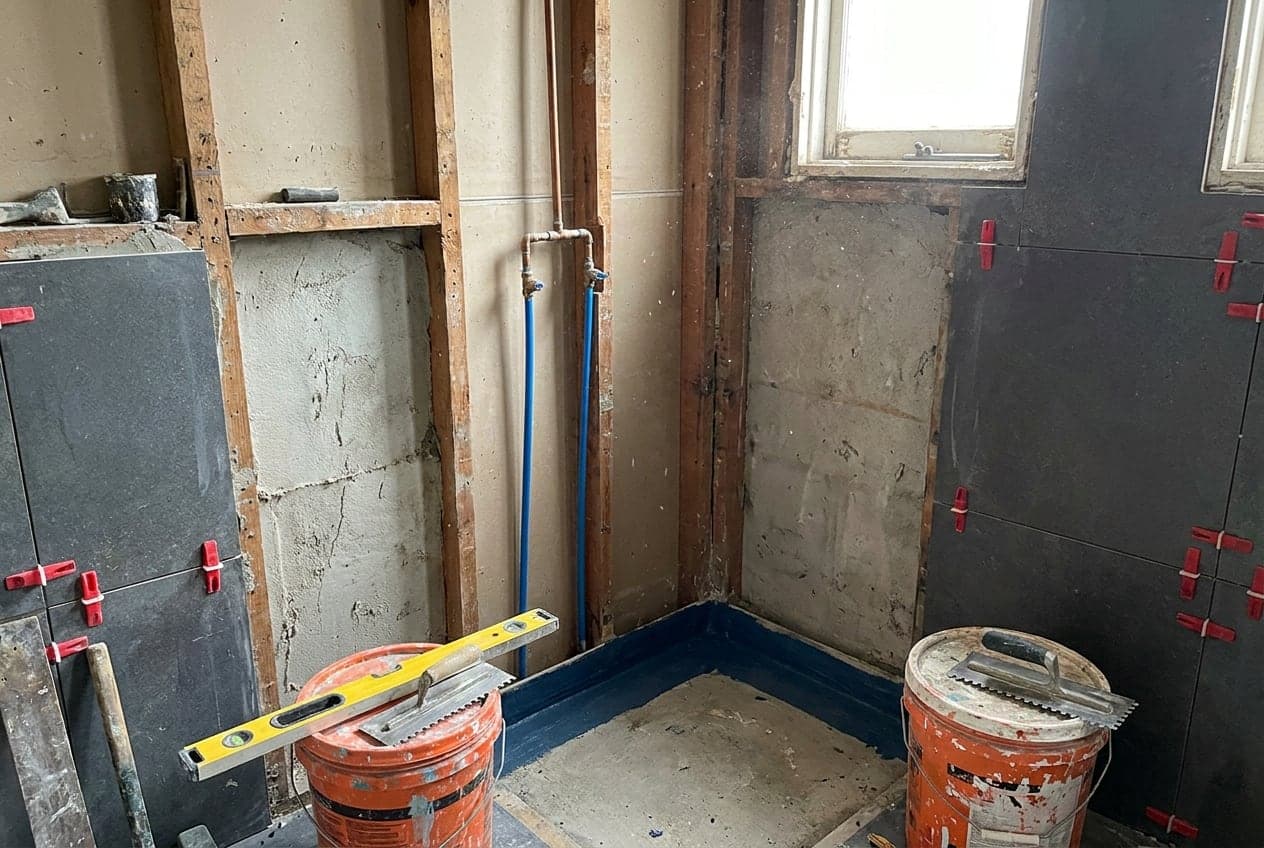 A bathroom stripped back to timber studs mid-renovation, with exposed pipework rough-in, large-format grey tiles going up, waterproofing membrane in the floor corner, and a spirit level and trowels resting on buckets